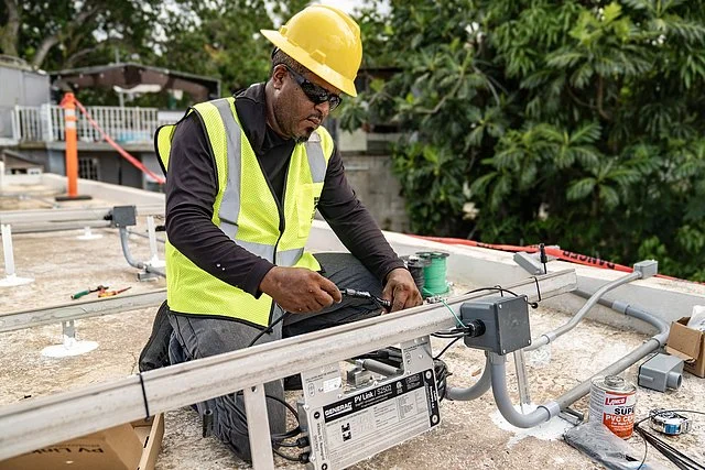 A construction worker in a yellow hard hat and reflective vest using a tool on a metal railing on a rooftop, with trees in the background.