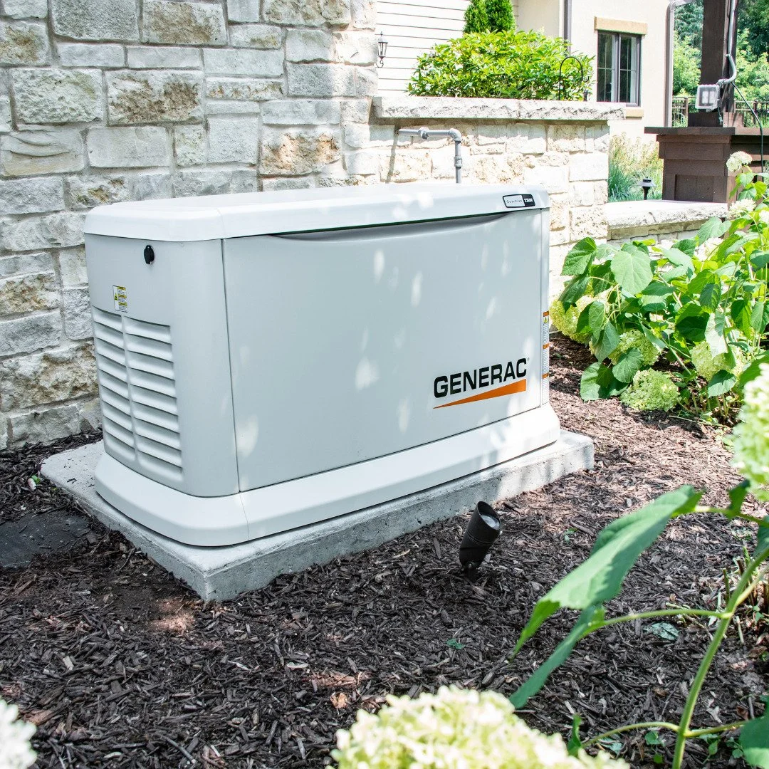 A Generac backup generator installed on a concrete pad next to a stone wall and garden plants outside a house.