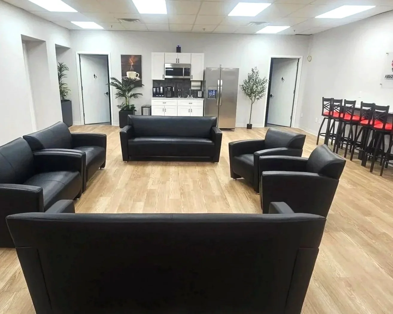 A lounge area with five black leather chairs arranged in a semi-circle around an empty space on a wooden floor, with a kitchenette in the background and a row of high chairs with red cushions on the right side.