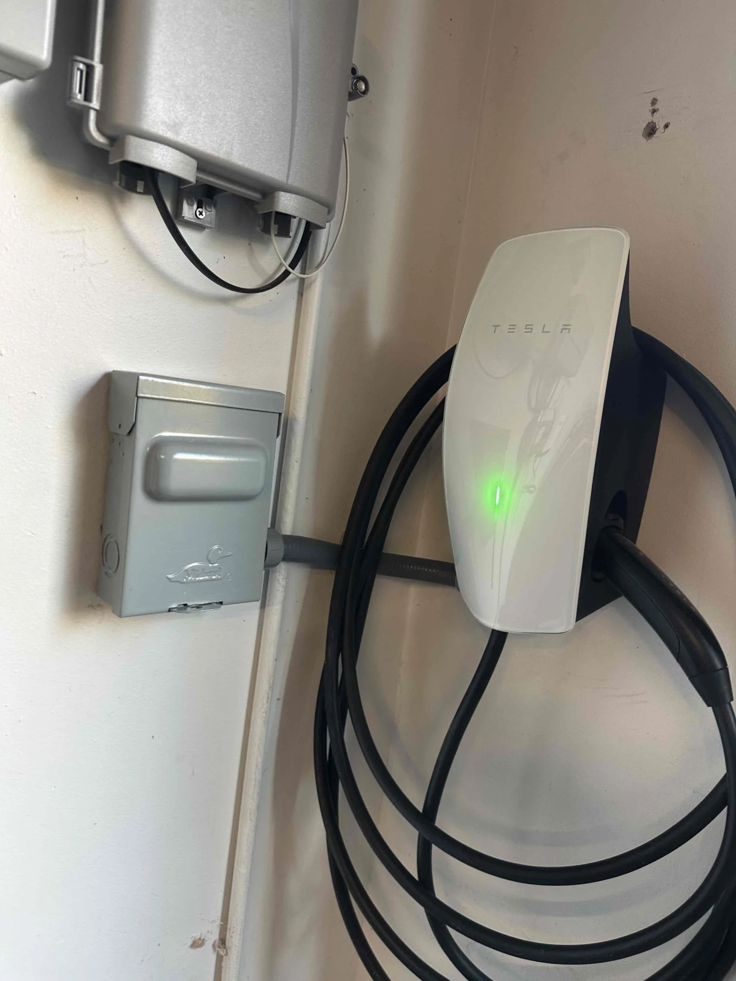 Wall with electrical outlets, a metal junction box, and a Tesla wall charger with cables connected.