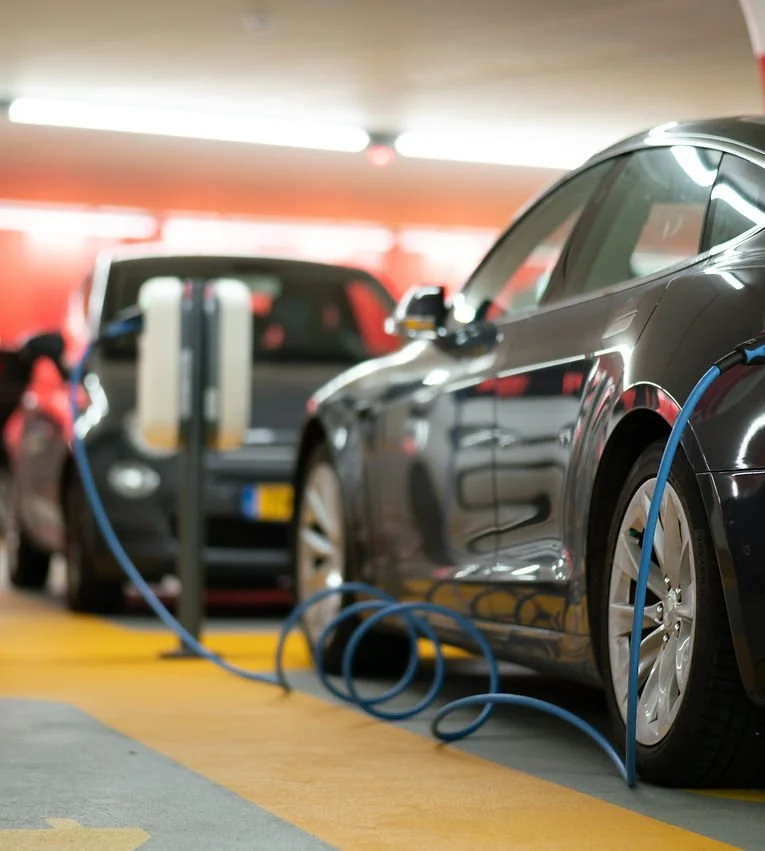 A black electric car plugged into a charging station in a parking garage, with another vehicle in the background.