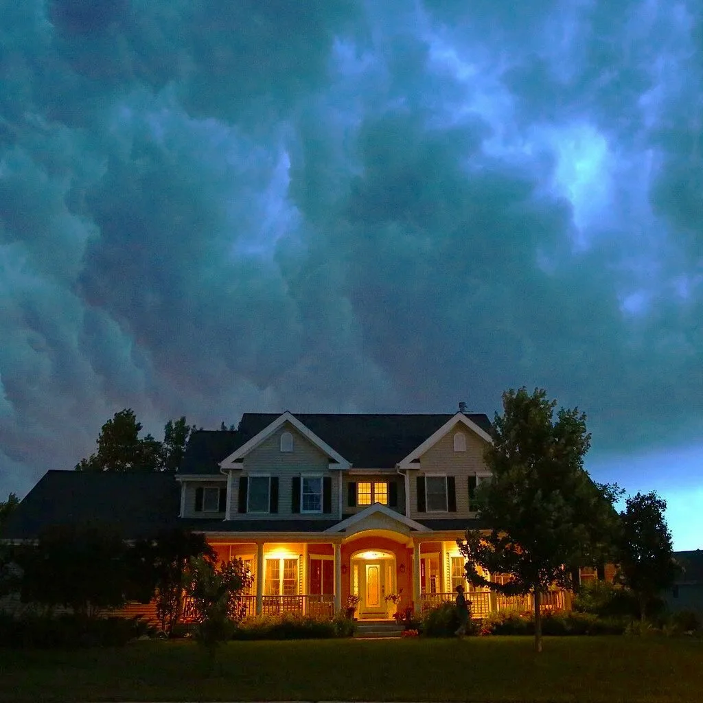 A large two-story house with lit windows and a front porch, set against a dark, cloudy sky at dusk.