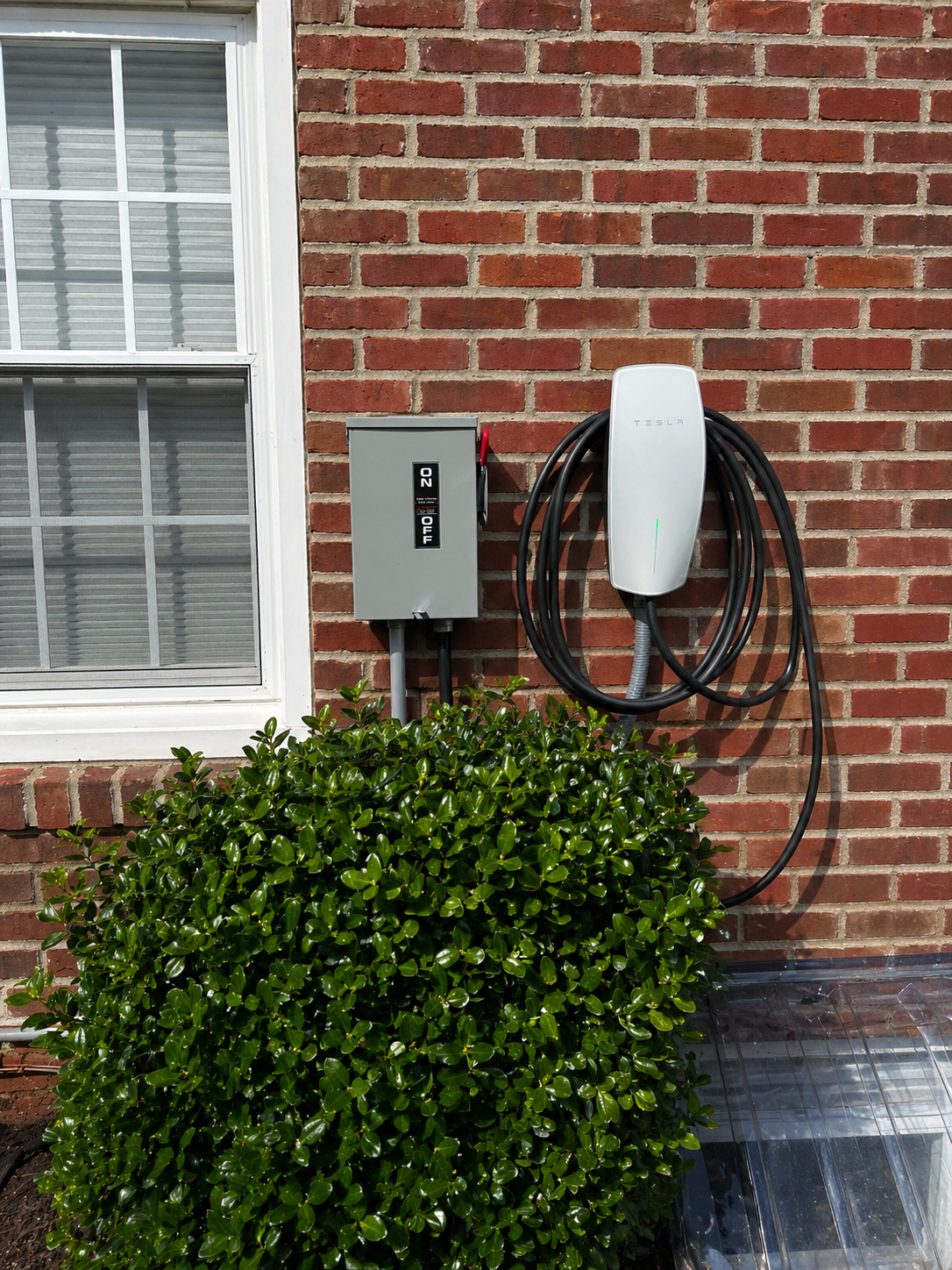 Electric vehicle charger mounted on a brick wall next to a gray electrical box and a window, with a green shrub in front.