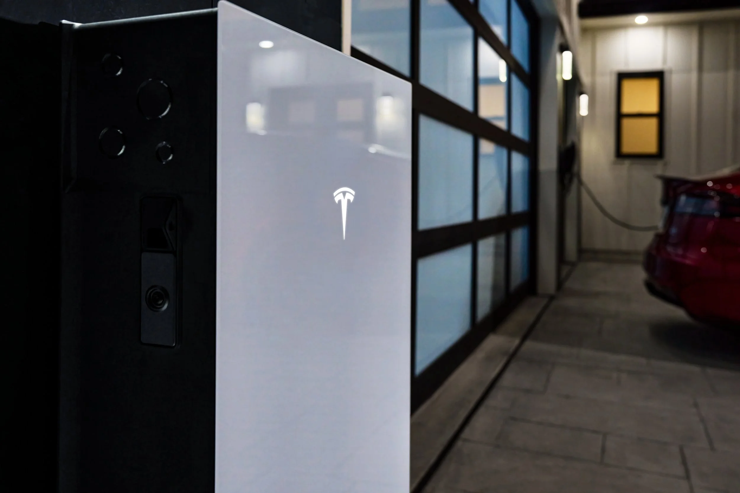 A Tesla electric vehicle charging station with a Tesla logo, located in an indoor parking garage, next to a red car and behind a glass wall.