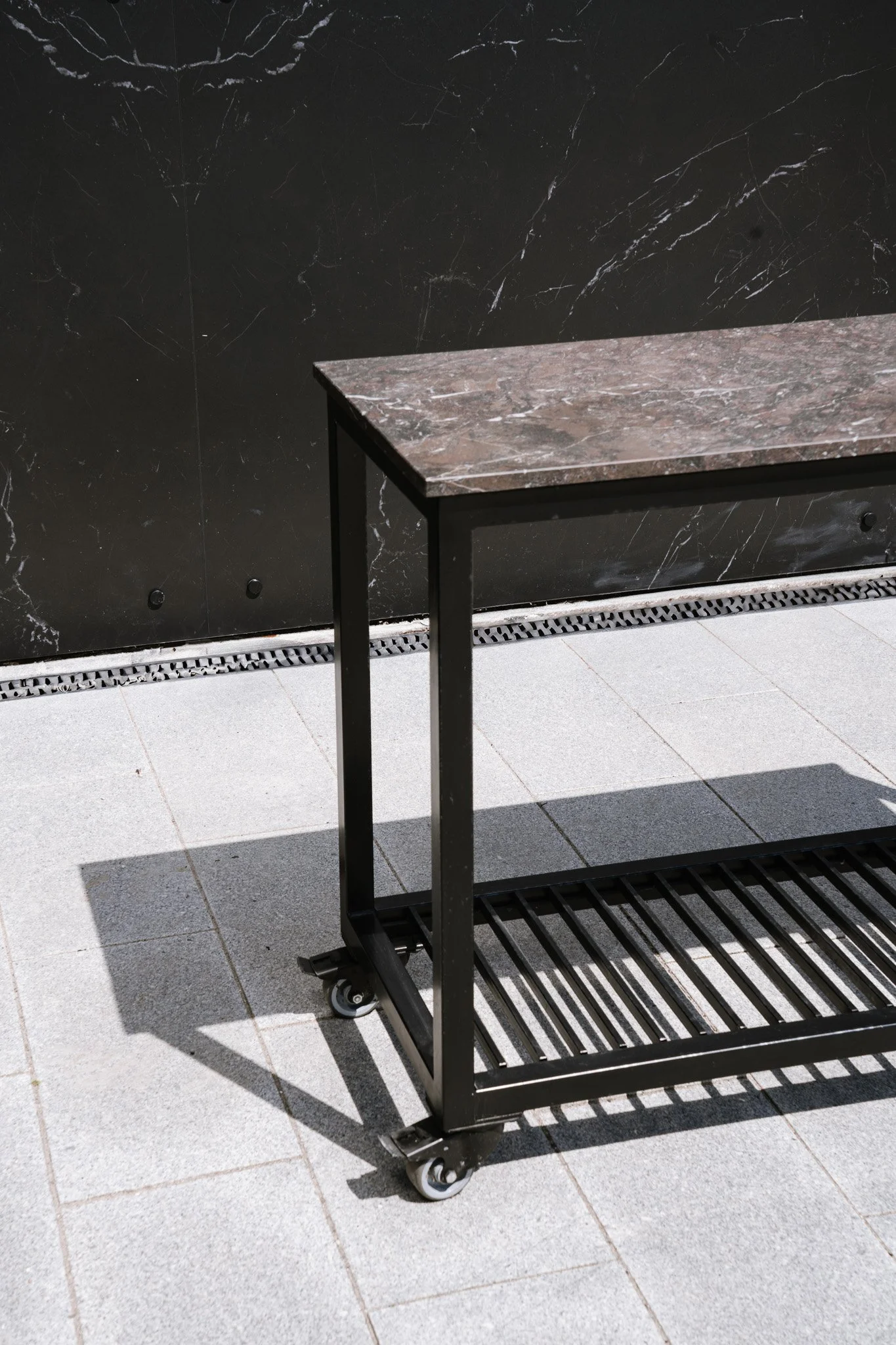 En sort metalkonstruktion med hjul og marmorbordplade, designet som en del af dit udekøkken. Placeret udendørs på en brolagt flade foran en mørk marmorvæg med en vask under.