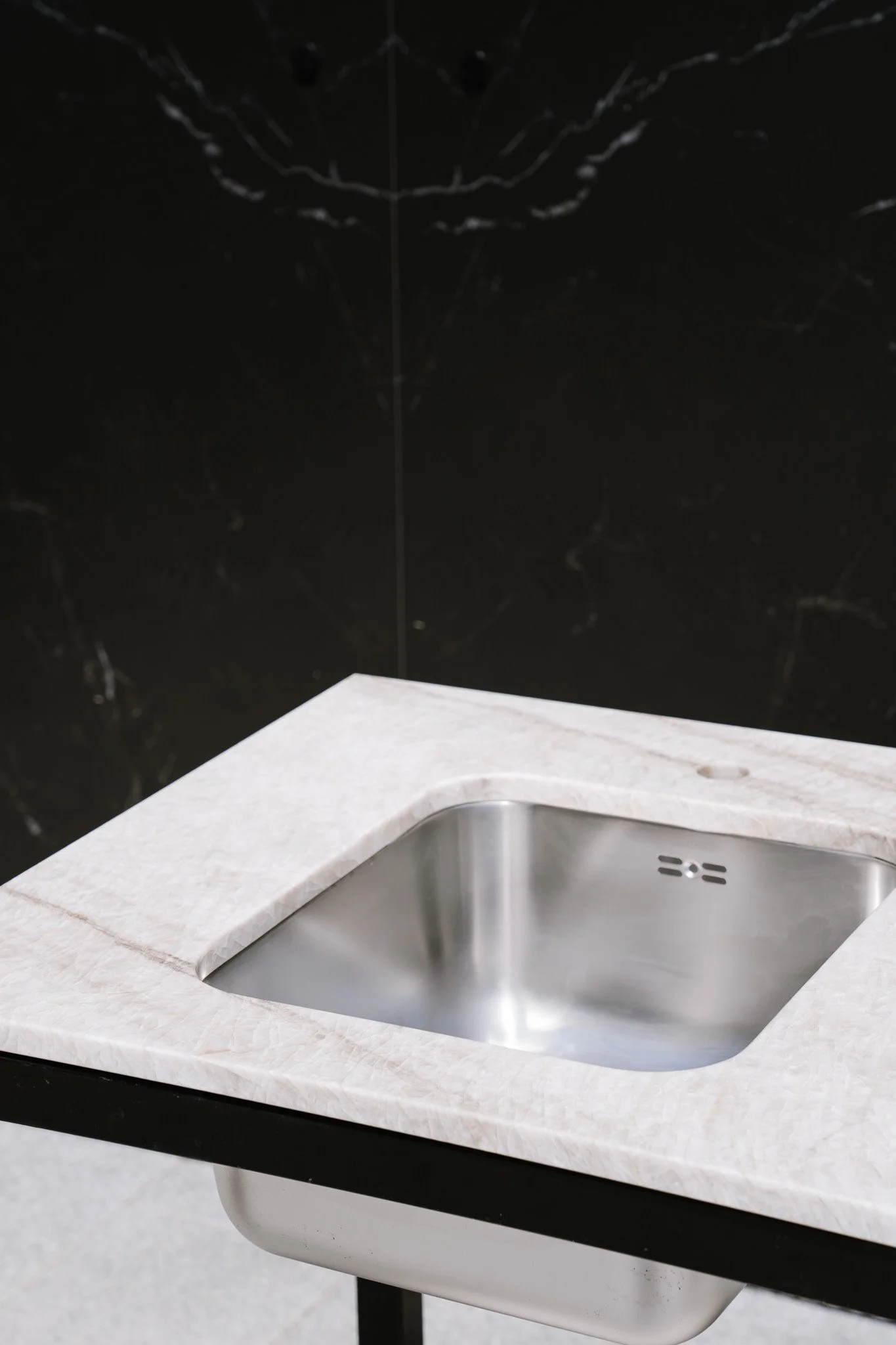 En sort metalkonstruktion med hjul og hvid marmorbordplade, designet som en del af dit udekøkken. Placeret udendørs på en brolagt flade foran en mørk marmorvæg med en rustfri stål vask under.