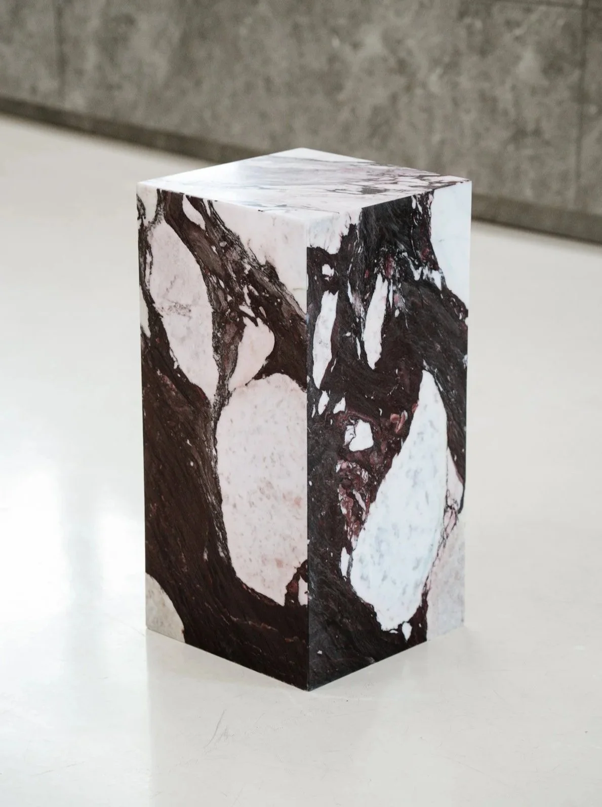 Et pedestal med marmormønster på overfladen, placeret på en hvid flade med grå baggrund. Kube piedestal i den smukke high-end marmor Calacatta Viola.