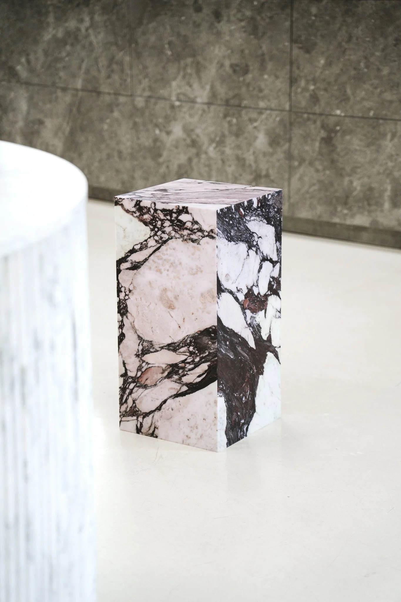 En pedestal med marmormønster på overfladen, placeret på en hvid flade med grå baggrund. Kube piedestal i den smukke high-end marmor Calacatta Viola.