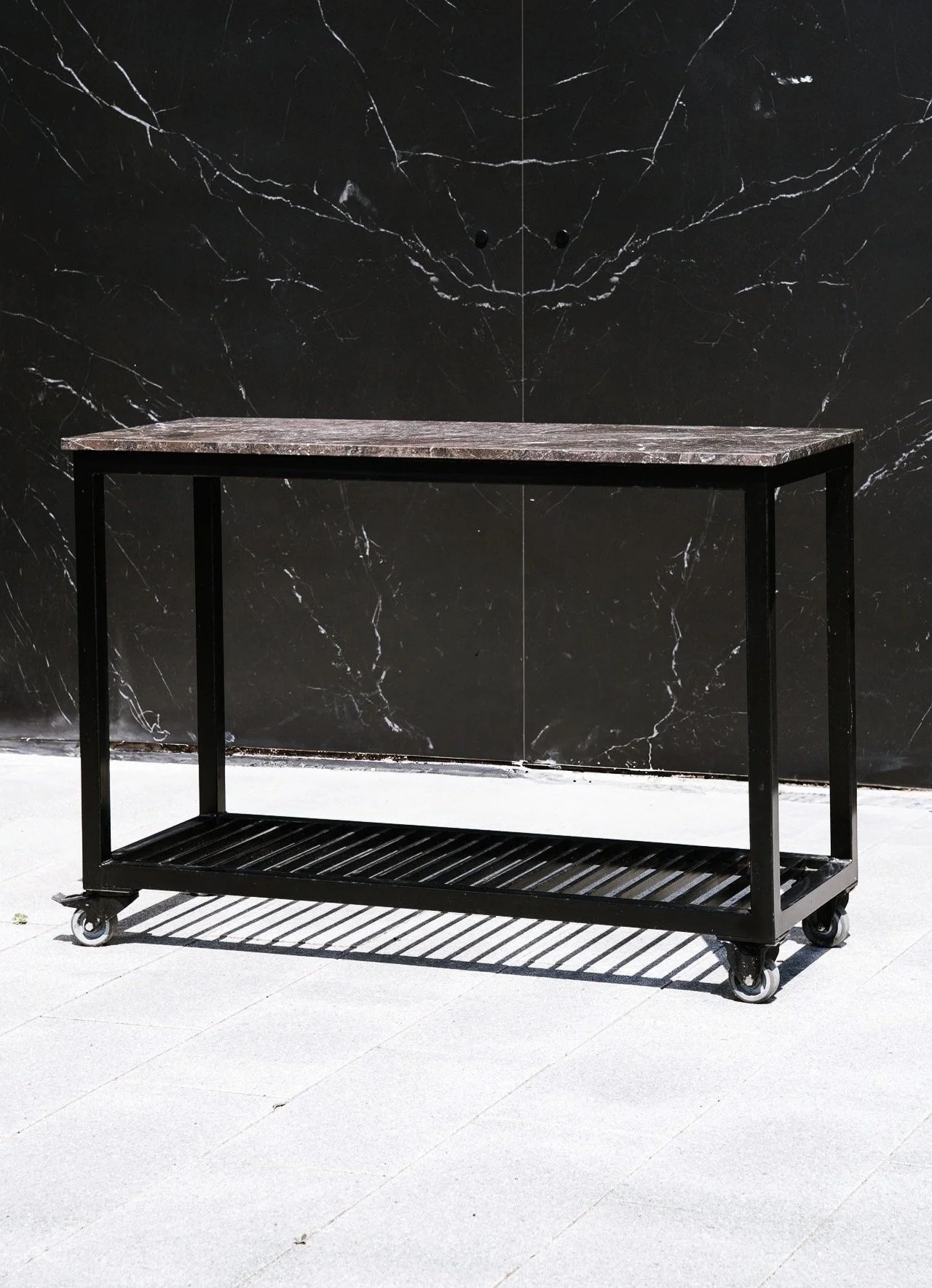 En sort metalkonstruktion med hjul og marmorbordplade, designet som en del af dit udekøkken. Placeret udendørs på en brolagt flade foran en mørk marmorvæg med en lille vask under.