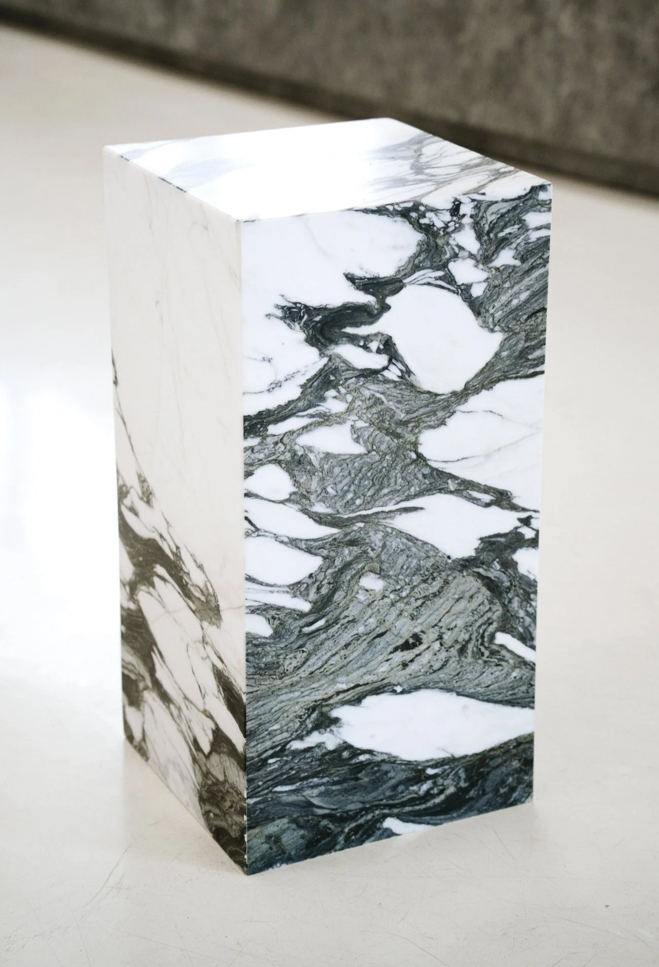 Et pedestal med sort-hvidt marmormønster på overfladen, placeret på en hvid flade med grå baggrund. Kubus piedestal i marmoren Verde Fantastico,