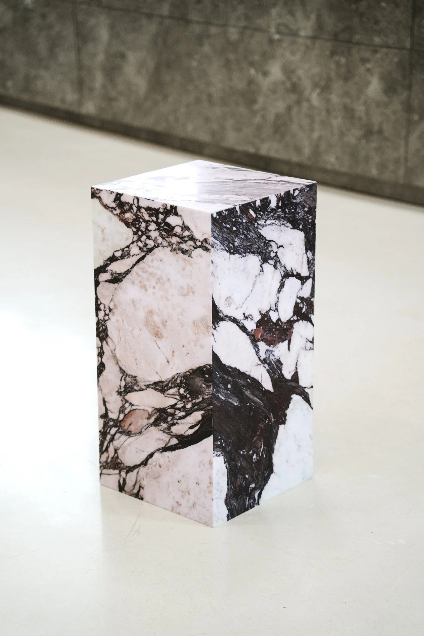 En pedestal med marmormønster på overfladen, placeret på en hvid flade med grå baggrund. Kube piedestal i den smukke high-end marmor Calacatta Viola.