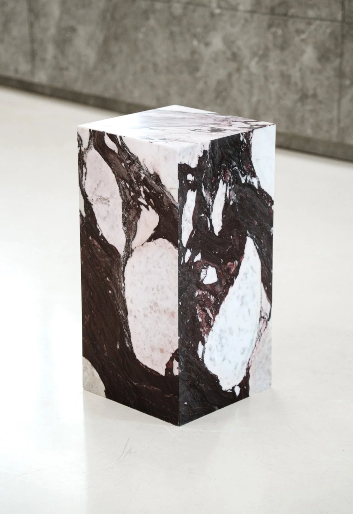 En pedestal med marmormønster på overfladen, placeret på en hvid flade med grå baggrund. Kube piedestal i den smukke high-end marmor Calacatta Viola.