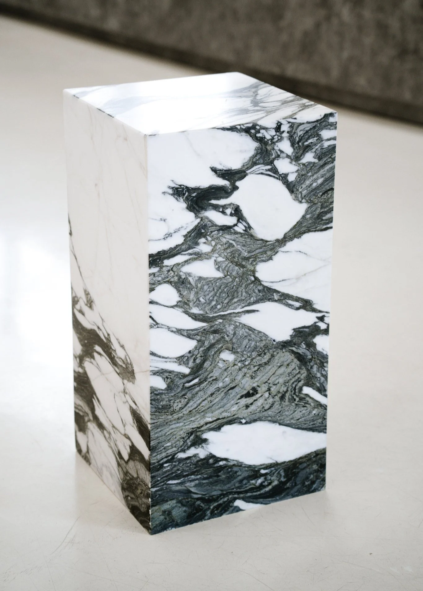 En pedestal med sort-hvidt marmormønster på overfladen, placeret på en hvid flade med grå baggrund. Kubus piedestal i marmoren Verde Fantastico.