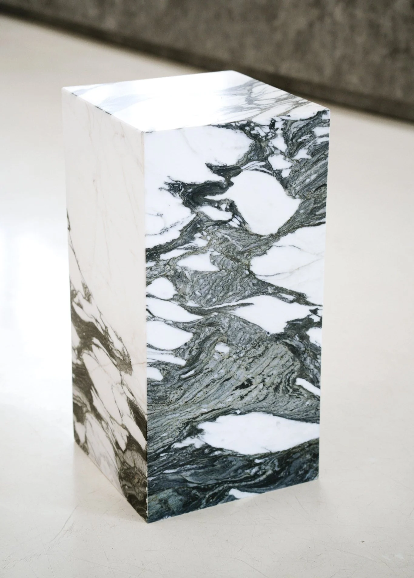 Et pedestal med sort-hvidt marmormønster på overfladen, placeret på en hvid flade med grå baggrund. Kubus piedestal i marmoren Verde Fantastico.