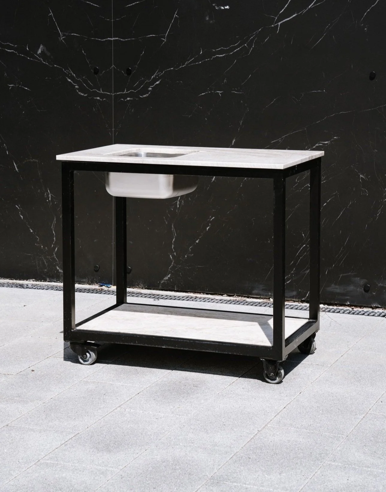 En vask monteret på en enkel metalkonstruktion med hjul og bordplade i marmor, placeret foran en sort marmorvæg på et betongulv.