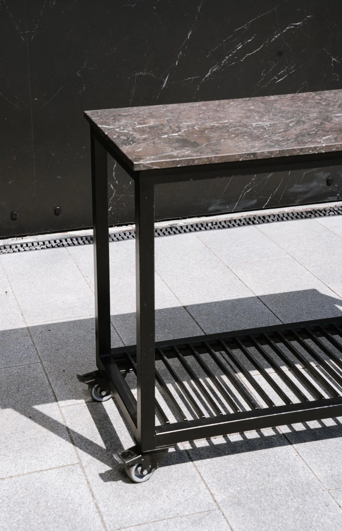 En sort metalkonstruktion med hjul og hvid marmorbordplade, designet som en del af dit udekøkken. Placeret udendørs på en brolagt flade foran en mørk marmorvæg med en lille vask under.