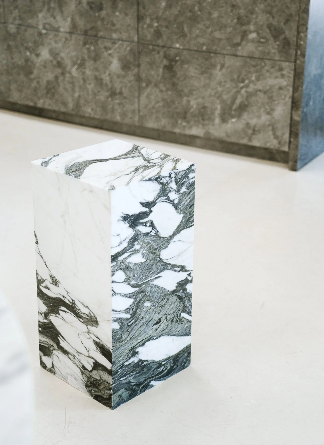 En pedestal med sort-hvidt marmormønster på overfladen, placeret på en hvid flade med grå baggrund. Kubus piedestal i marmoren Verde Fantastico.