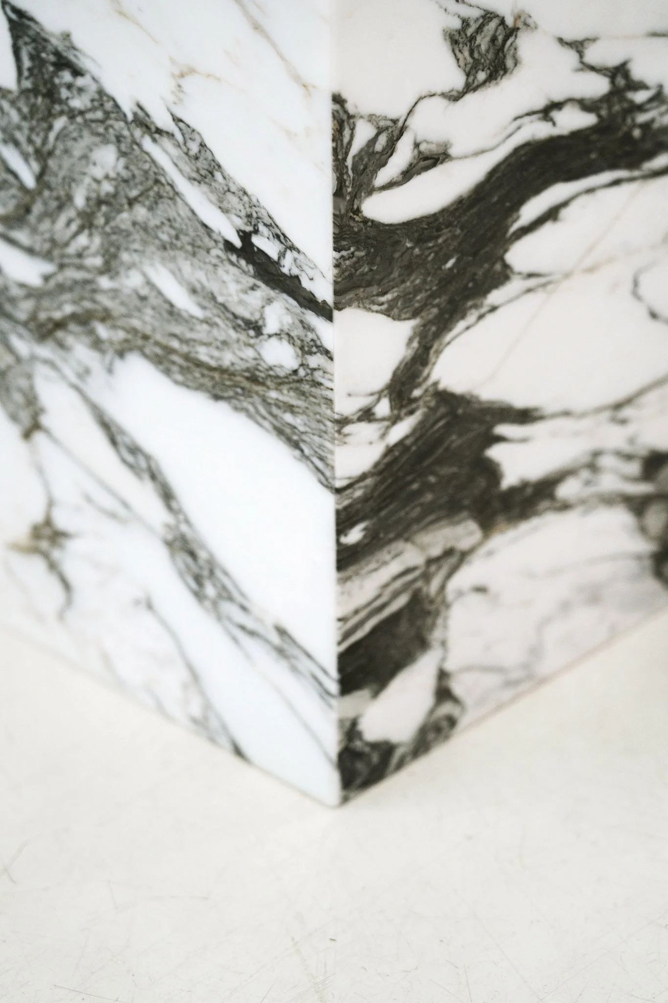 En pedestal med sort-hvidt marmormønster på overfladen, placeret på en hvid flade med grå baggrund. Kubus piedestal i marmoren Verde Fantastico.