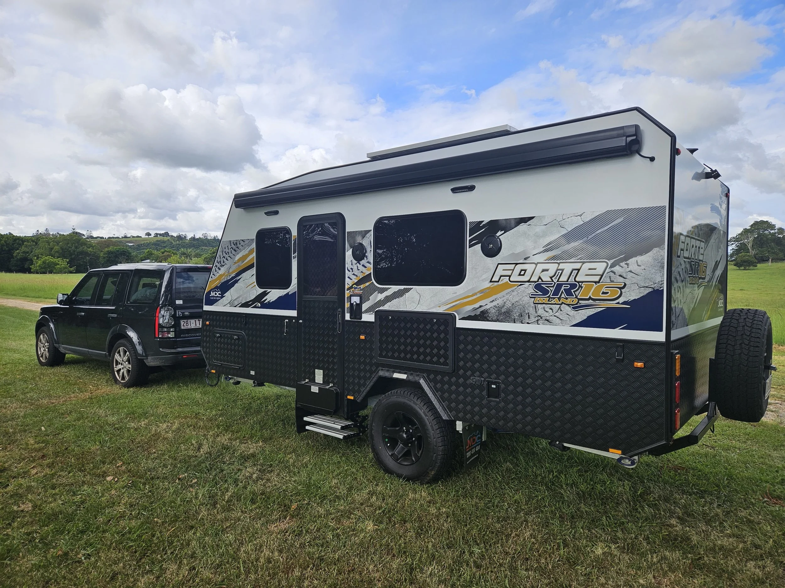 A black SUV towing a large, black and white off-road camper trailer with graphics reading Forte SR16 Island, parked on grass in a rural area with green fields and trees, under a partly cloudy sky.