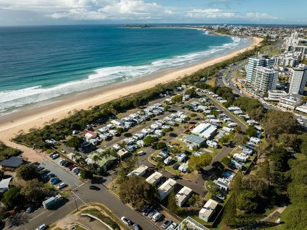 Aerial view of a coastal area with a sandy beach, ocean waves, and a seaside town with buildings and a parking lot.