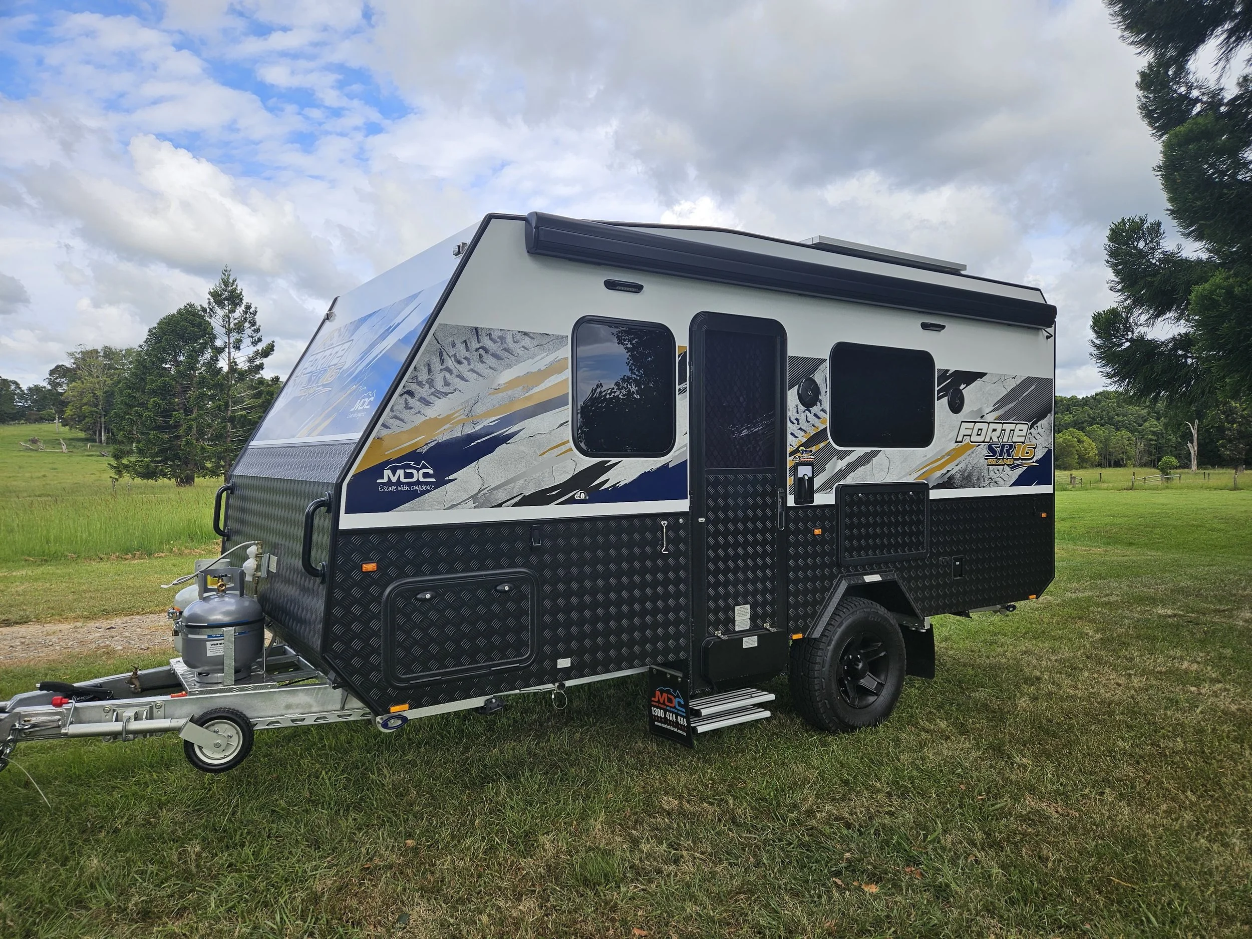 A modern travel trailer with black, white, and yellow graphics parked on grass with trees and cloudy sky in the background.