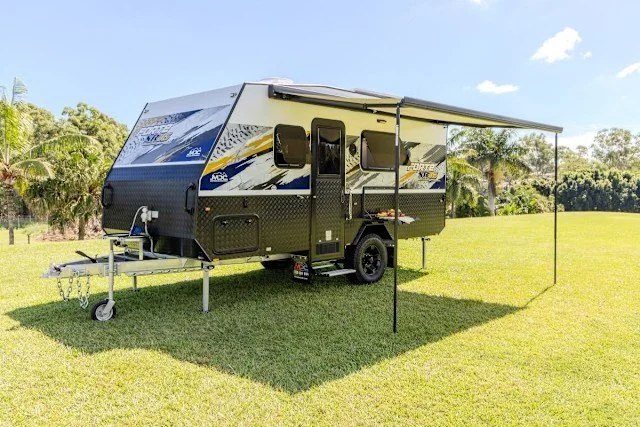 MDC Caravan with an extended awning and a tow hitch, parked on grassy field with palm trees and blue sky in the background.