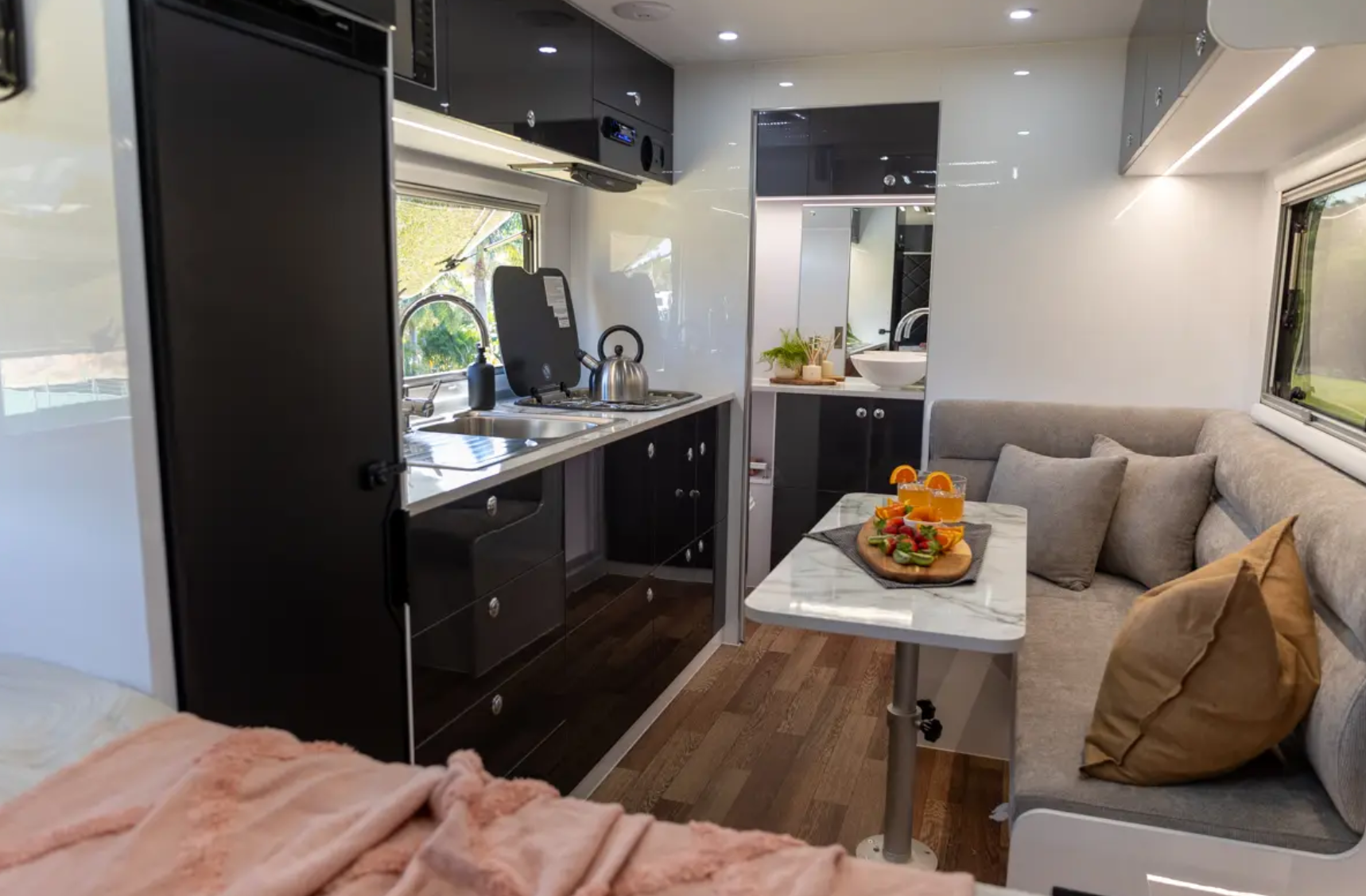 Interior of a modern RV with a compact kitchen area featuring black cabinets, a small sink, a stovetop with a kettle, a window, and a seating area with a table, which has a platter of snacks and drinks, all with natural light coming through the windows.