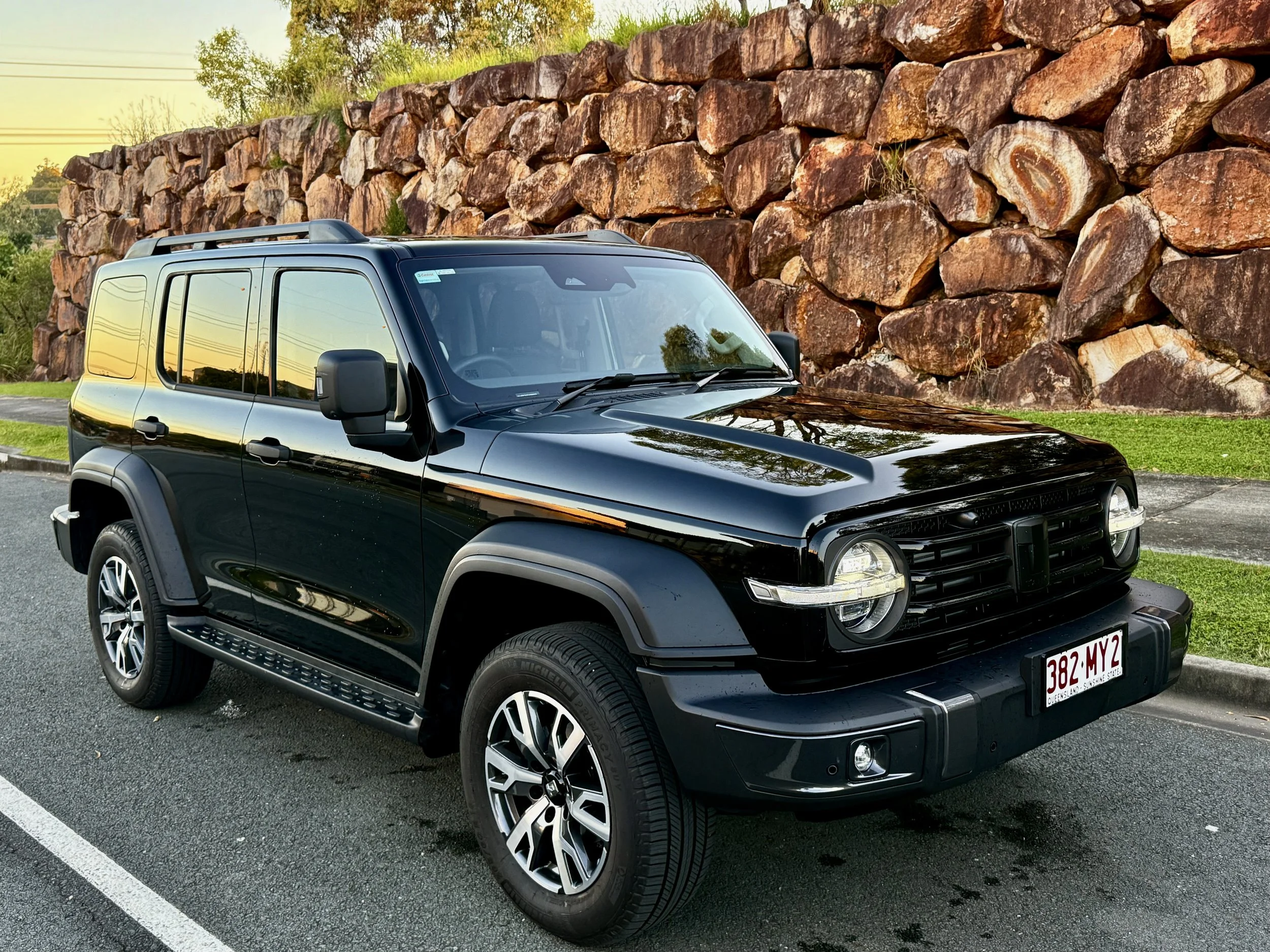 Black SUV parked on the street with a large rock wall and green grass in the background during sunset.