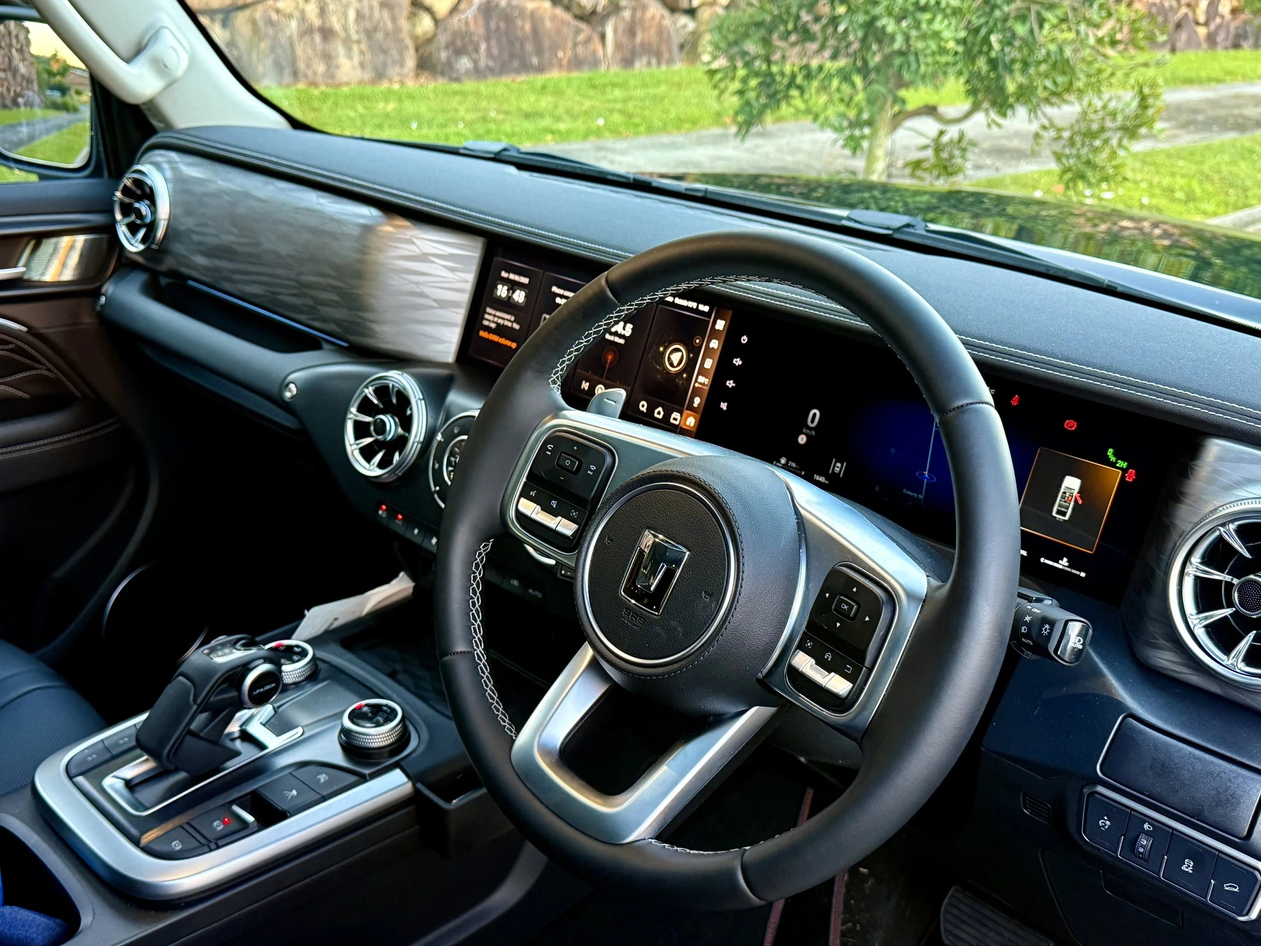 Interior view of a modern vehicle dashboard featuring a digital display, air vents, and gear shifter, with a lush green outdoor scene visible through the windshield.
