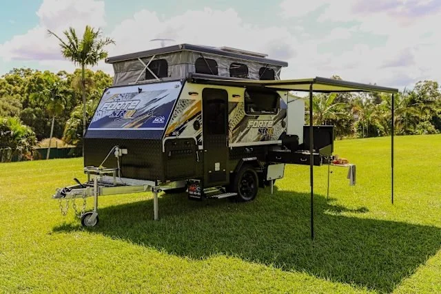 A compact travel trailer with a pop-up tent and an extended awning, set up on a grassy field with trees and cloudy sky in the background.