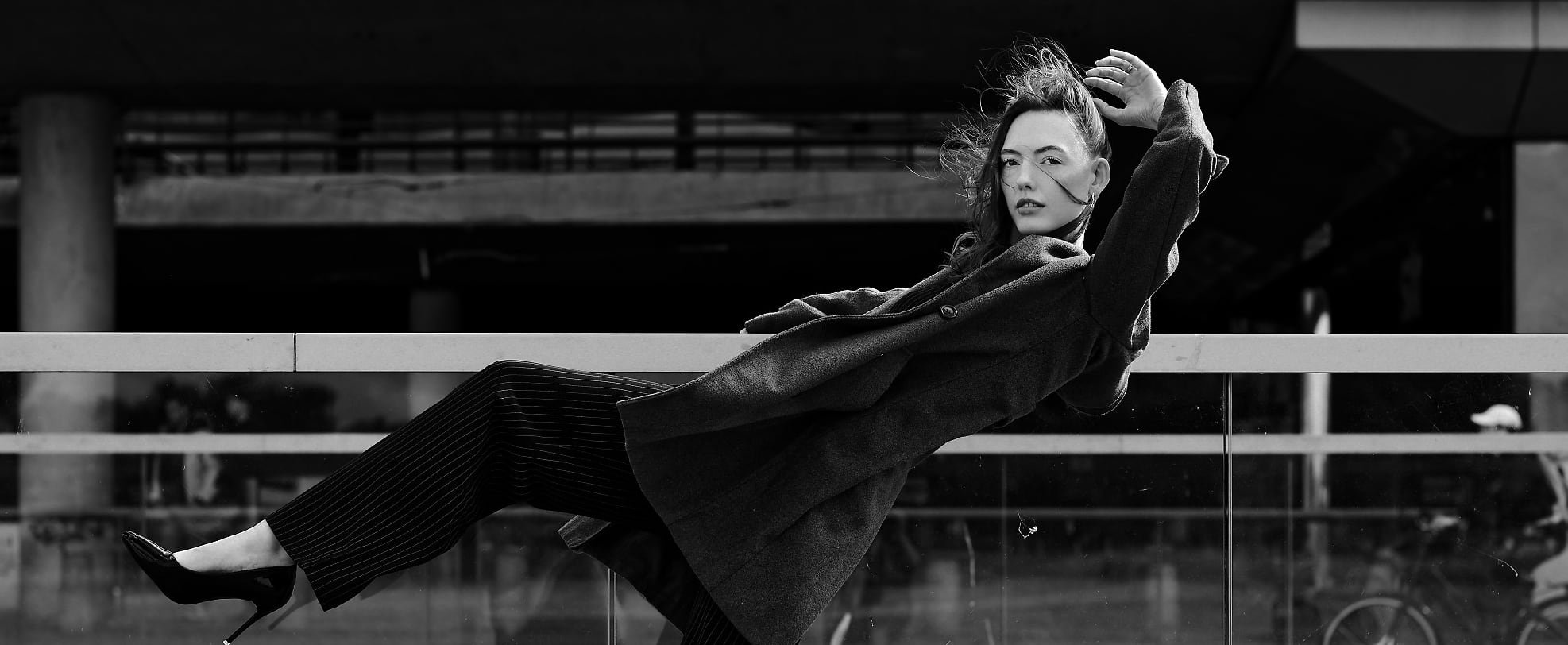 Black-and-white photo of a person in a dramatic pose, wearing a long coat and pinstripe pants, balancing with one leg raised against a glass railing.