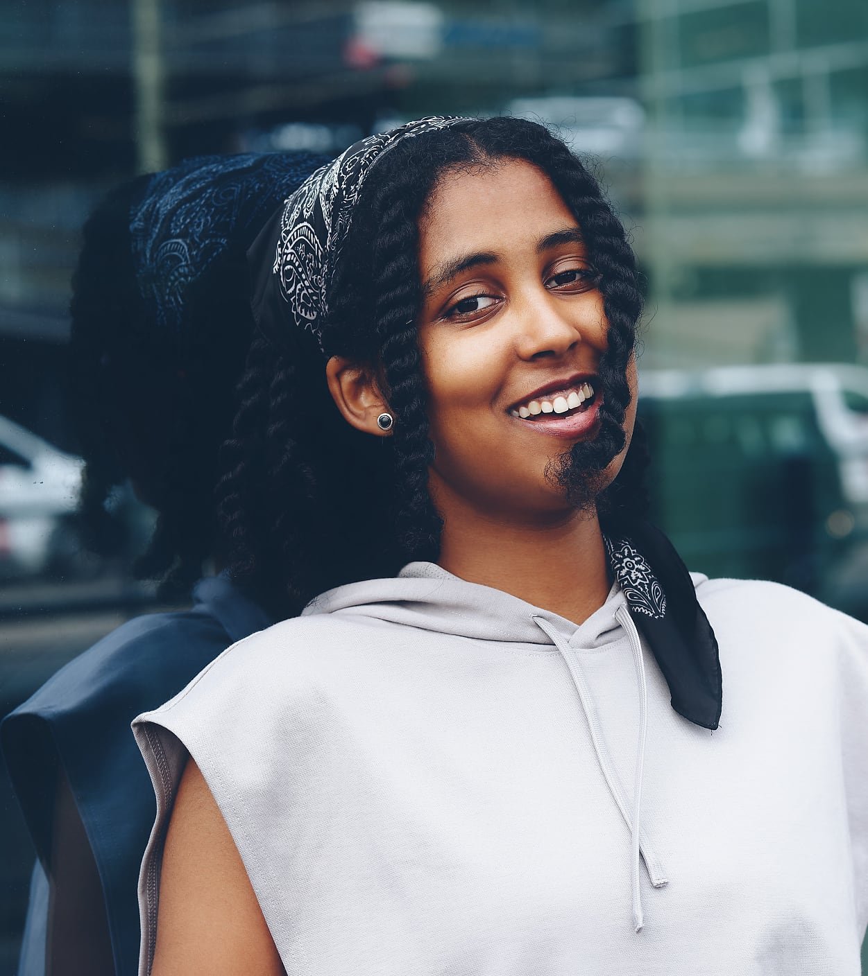A person with curly hair styled in braids and a patterned bandana is wearing a sleeveless light hoodie, standing in front of a reflective glass wall.