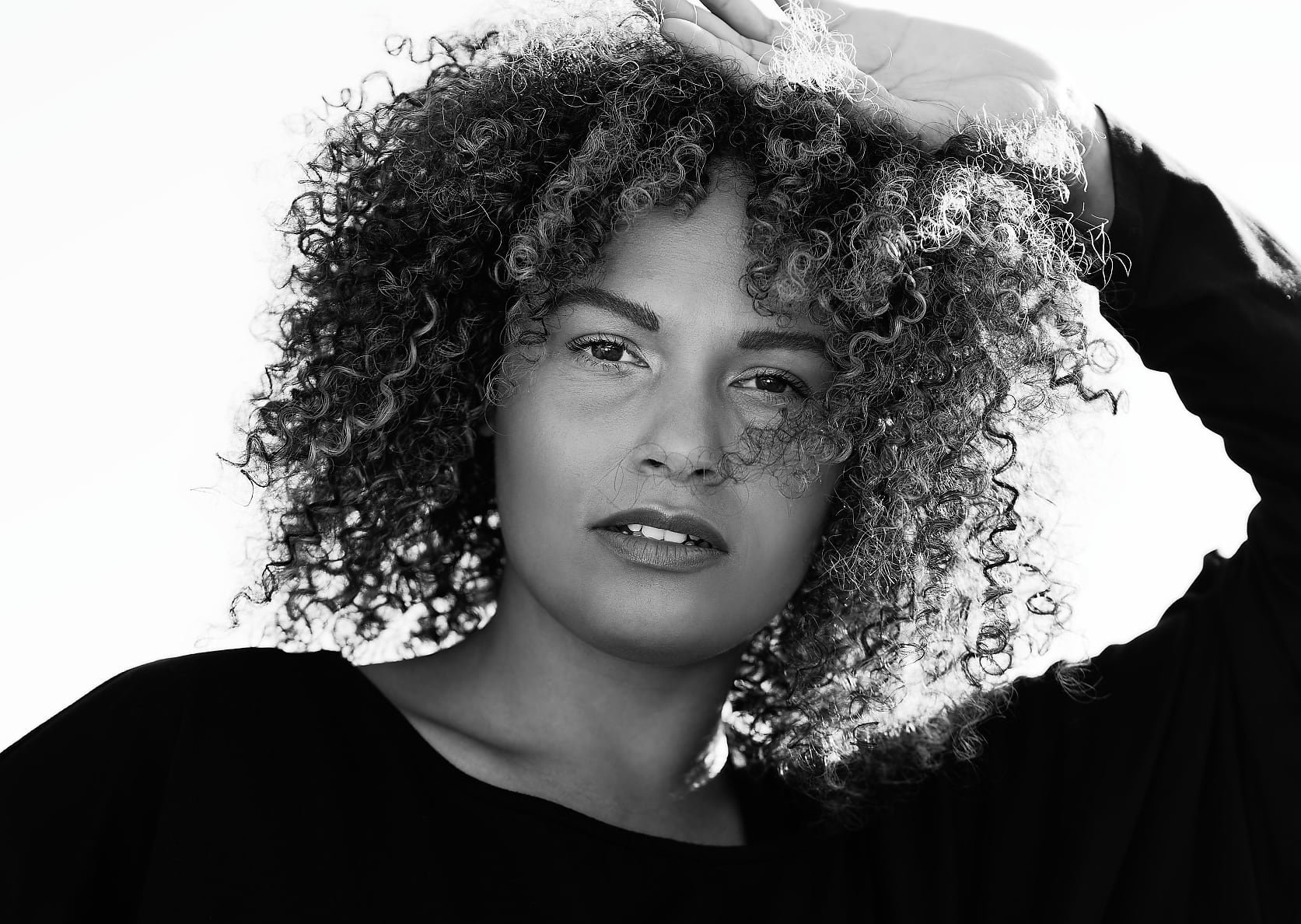 A person with voluminous, curly hair is standing against a bright background. They are wearing a loose, long-sleeved black top, and one arm is raised above their head, casting a shadow.