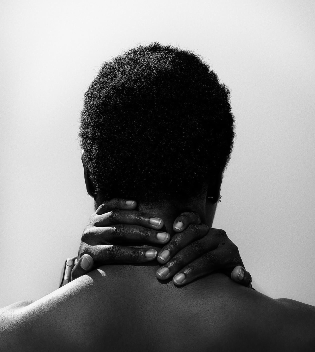 A black-and-white image captures the back of a person with short, natural, tightly coiled hair. Their hands are clasped around the back of their neck, and their bare shoulders are visible.