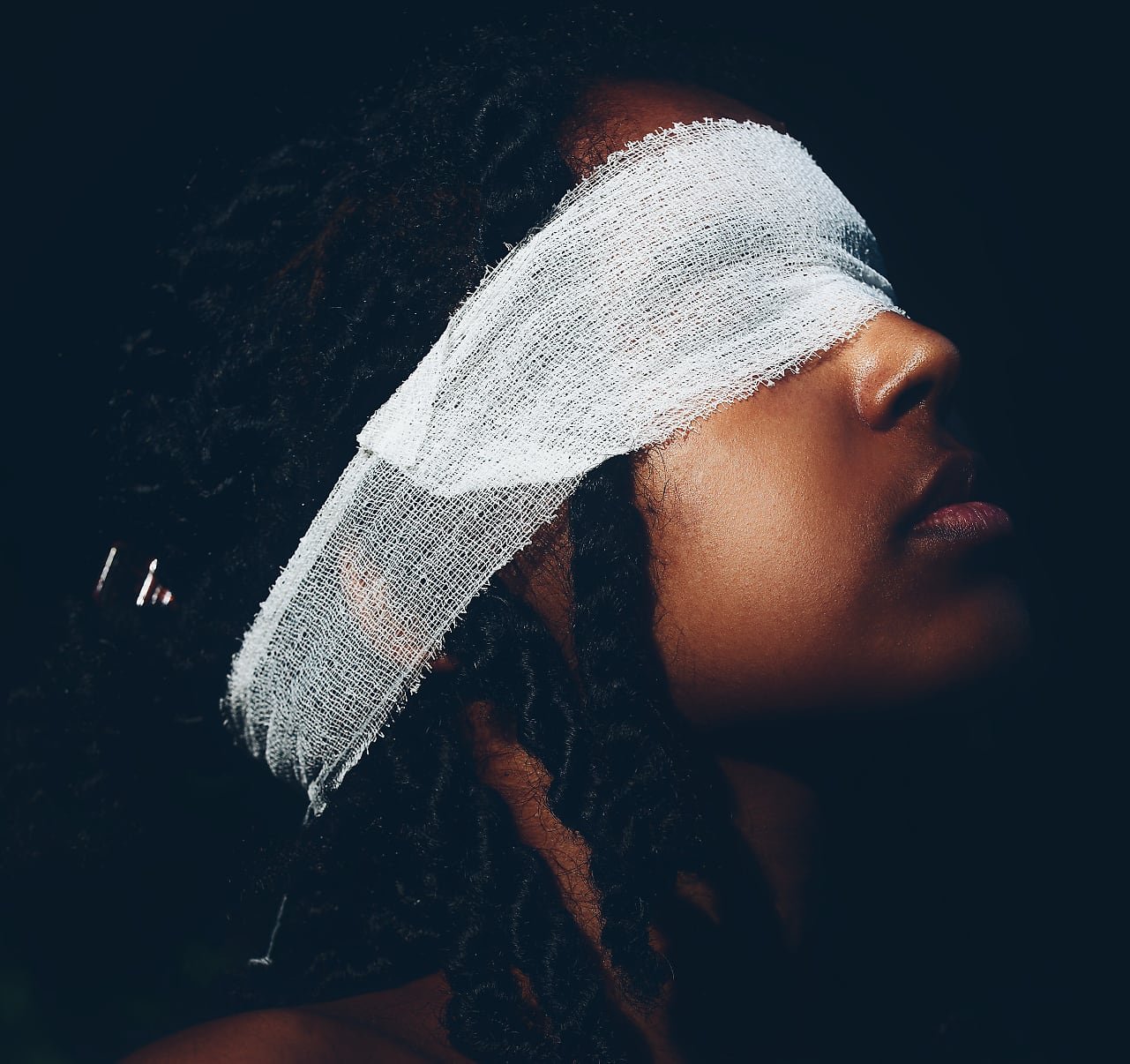 Artistic portrait of a person with textured hair, wearing gauze bandages over their eyes and arms, highlighted by dramatic lighting.