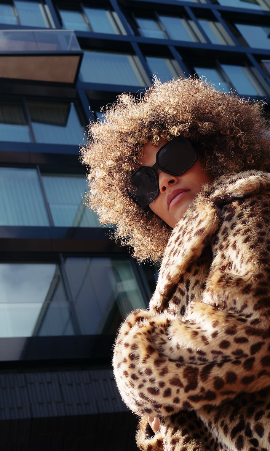 Low-angle shot of a person in a leopard print faux fur coat, standing in front of a modern glass building, hair illuminated by sunlight.