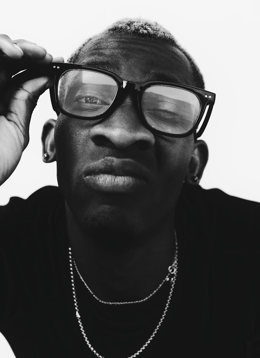 A person with short, light blond, tightly coiled hair is wearing dark glasses and layered chain necklaces. The photo is in black and white, and they are holding the glasses with one hand.