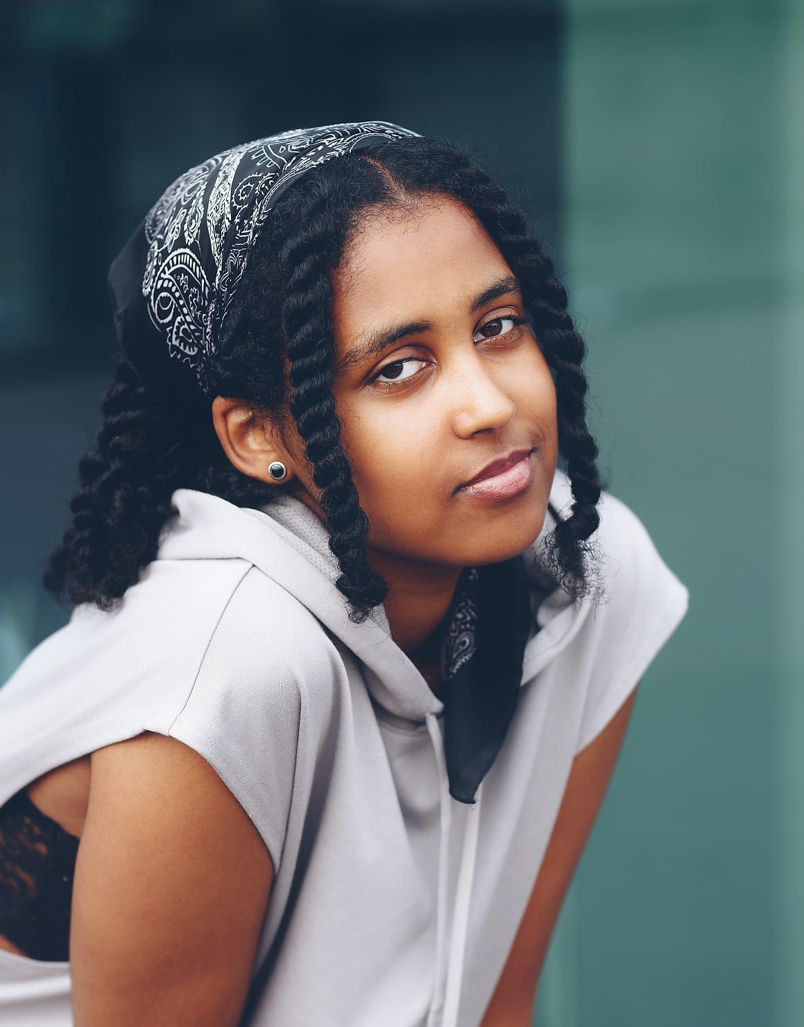 A person with curly hair in braids and a patterned bandana is wearing a sleeveless light hoodie, leaning forward in an urban environment.