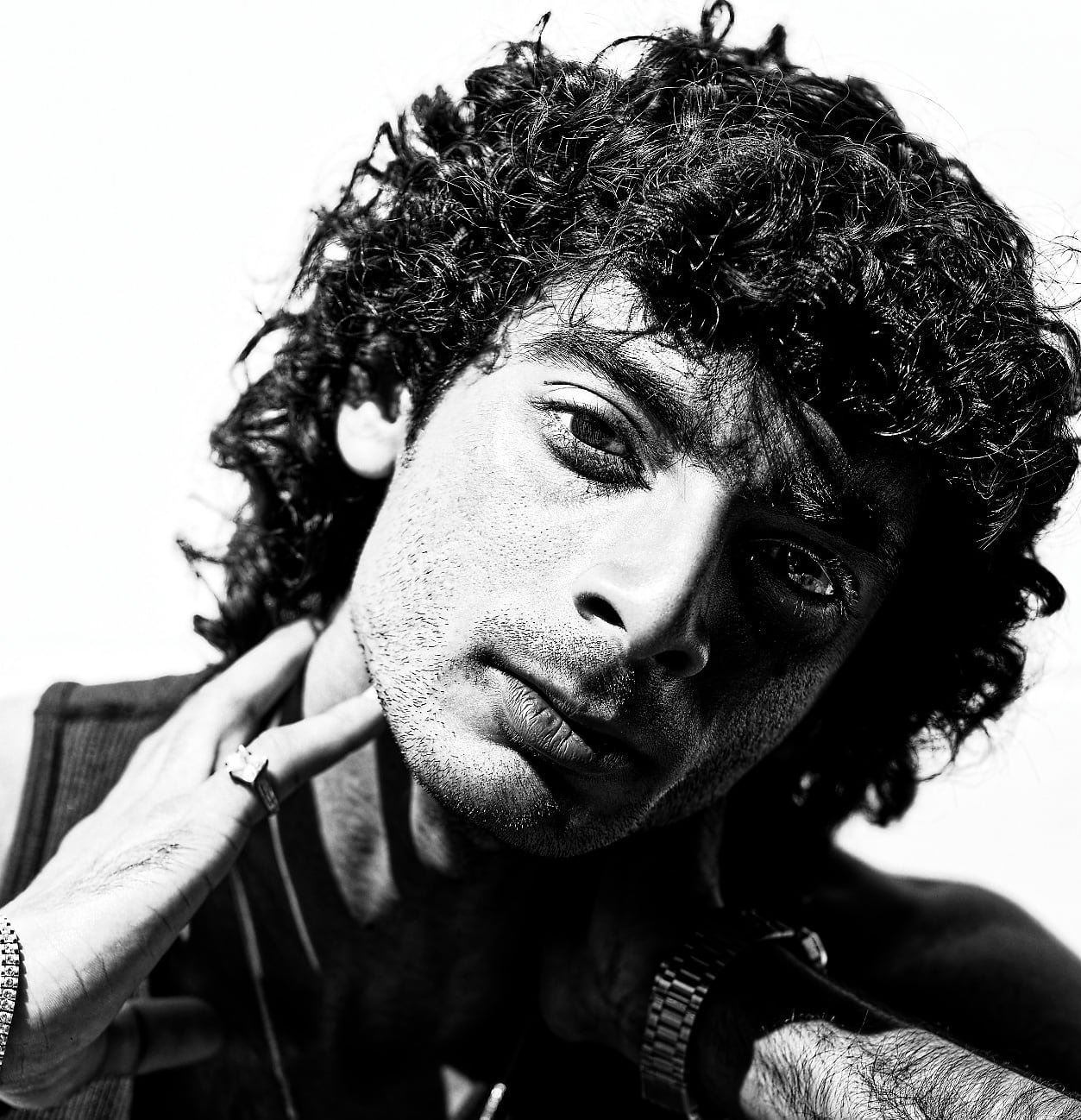 A black-and-white portrait of a person with thick, curly hair. They are wearing a watch and a ring, with one hand raised near their neck and the other partially visible. The background is bright and uncluttered, emphasizing the subject’s hair and acc