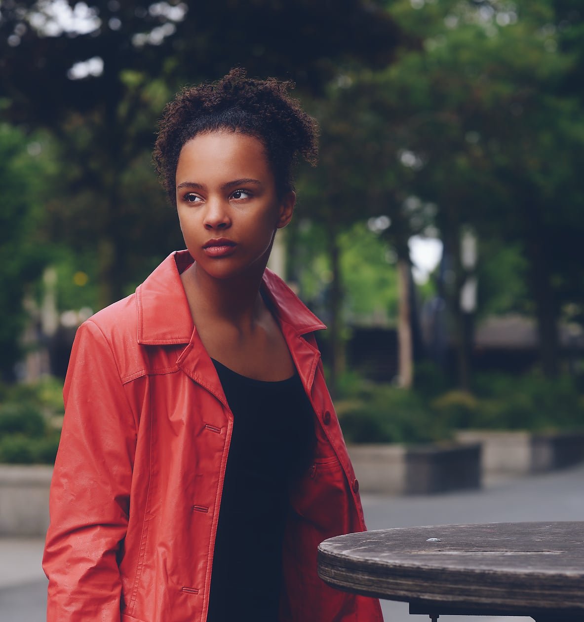 A person with curly hair in a coral trench coat and black top stands beside a round table in an outdoor urban park.