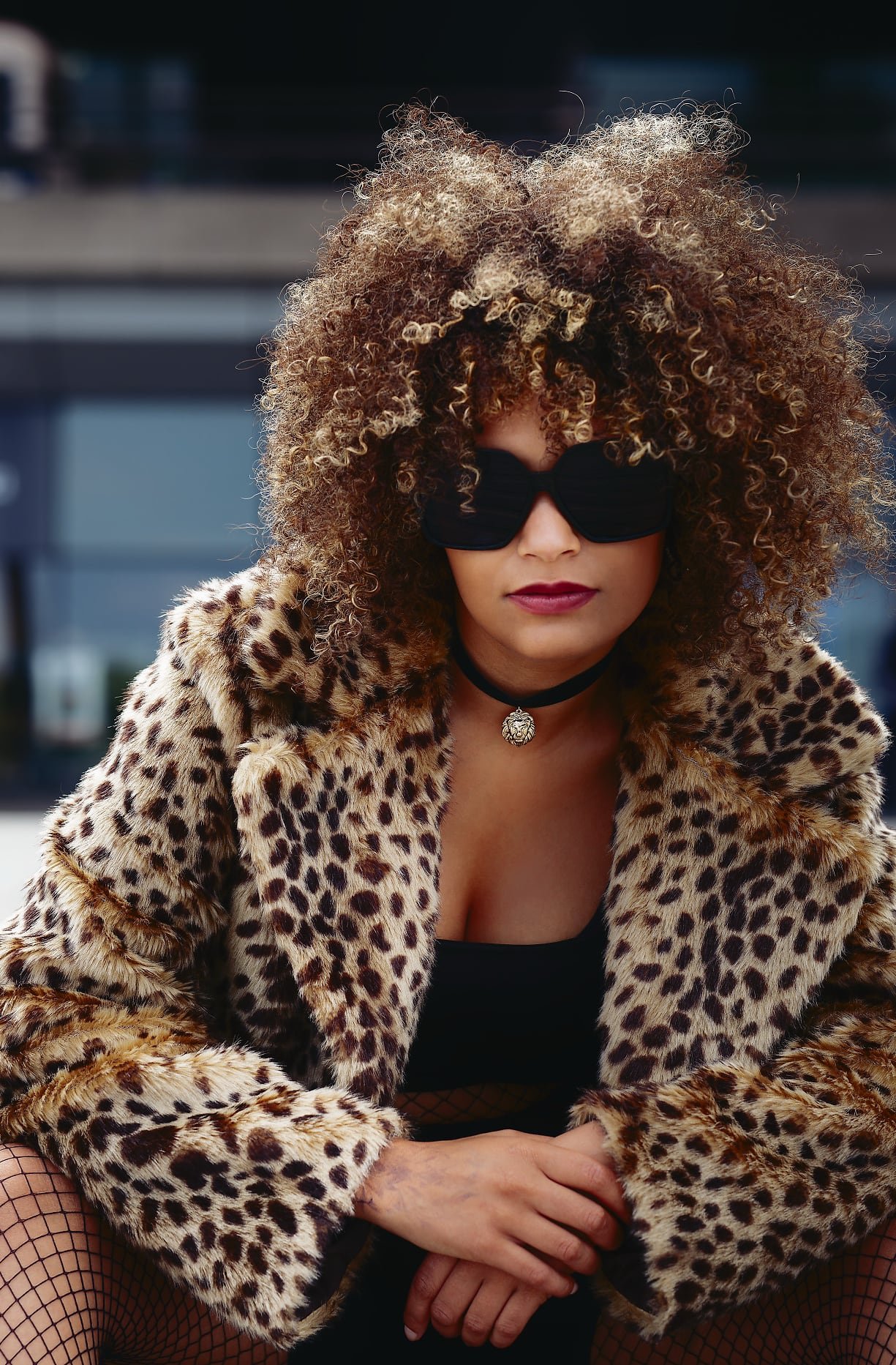 Fashion-forward image of a person in a leopard print faux fur coat, black top, and fishnet tights, sitting confidently in an urban setting.