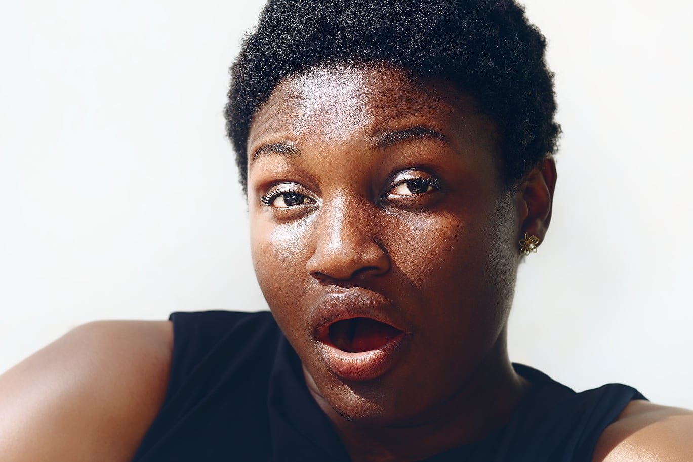 A person with short, natural, tightly coiled hair is sitting against a light background. They are wearing a sleeveless black top, and both arms are visible, showing a relaxed pose.