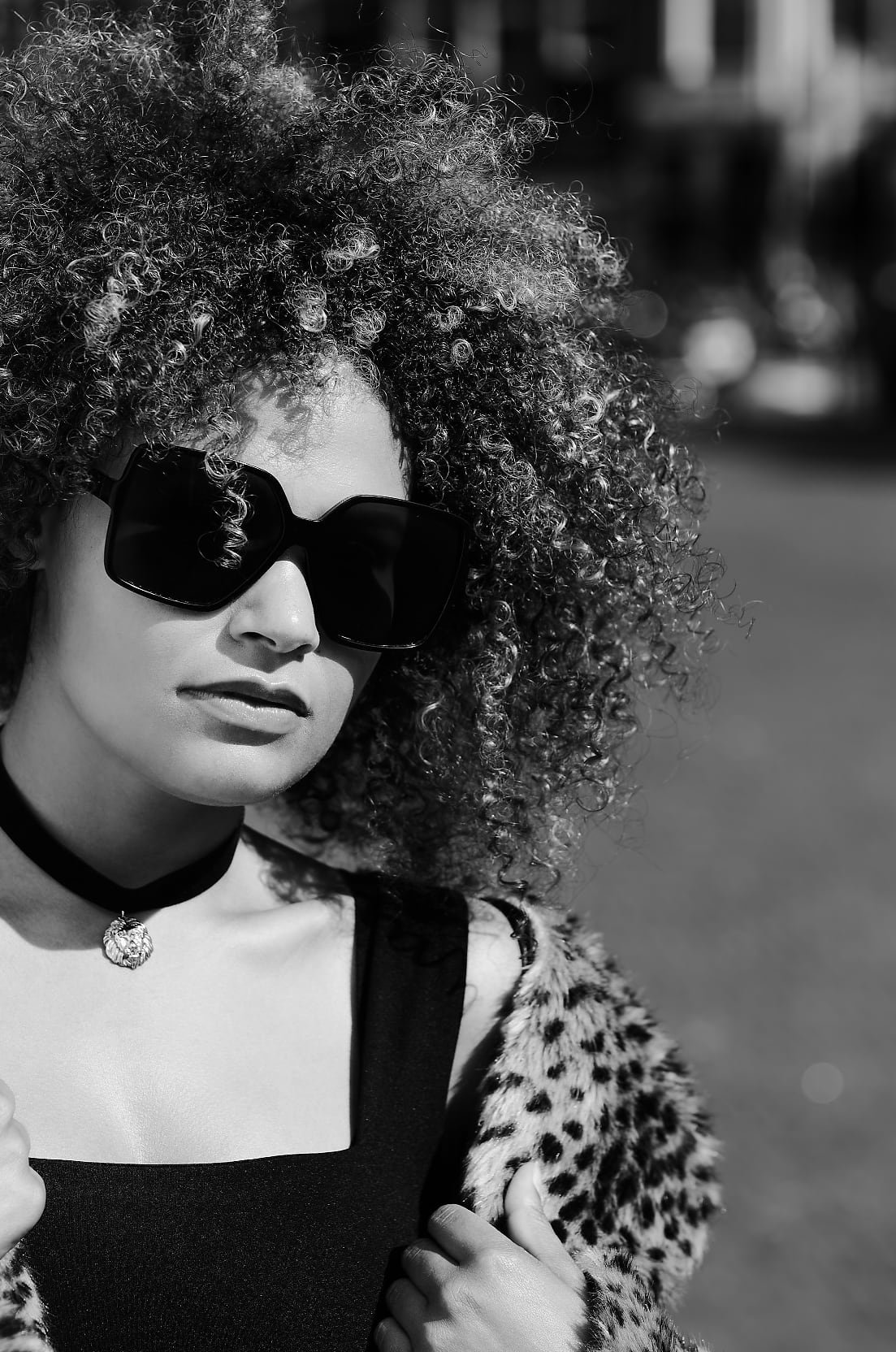 A person with voluminous curly hair is dressed in a leopard print coat, black top, and choker necklace, photographed outdoors in black and white.
