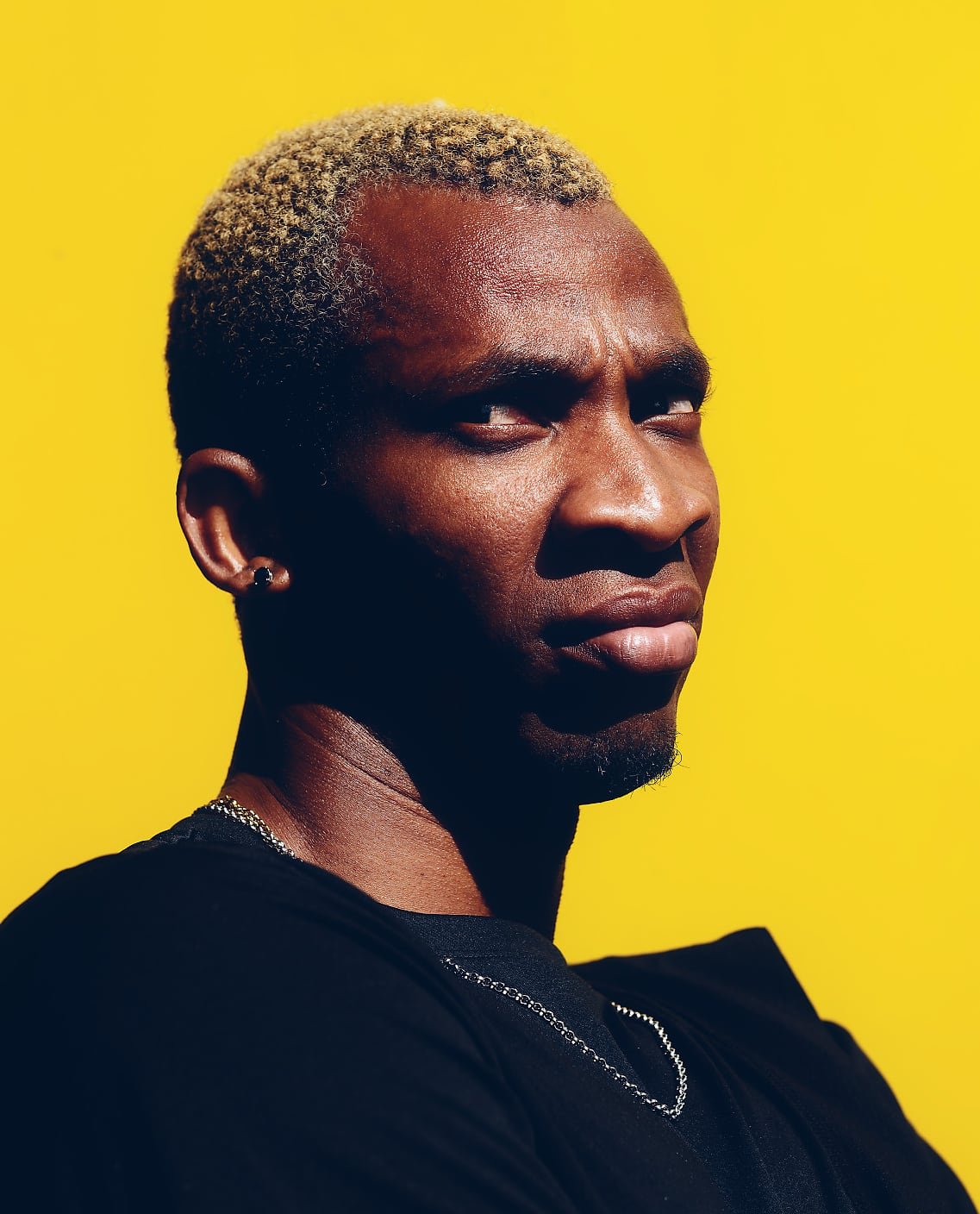 A person with short, tightly coiled hair dyed light blond is photographed against a bright yellow background. They are wearing a black T-shirt and a chain necklace, with one ear and side of the face visible.