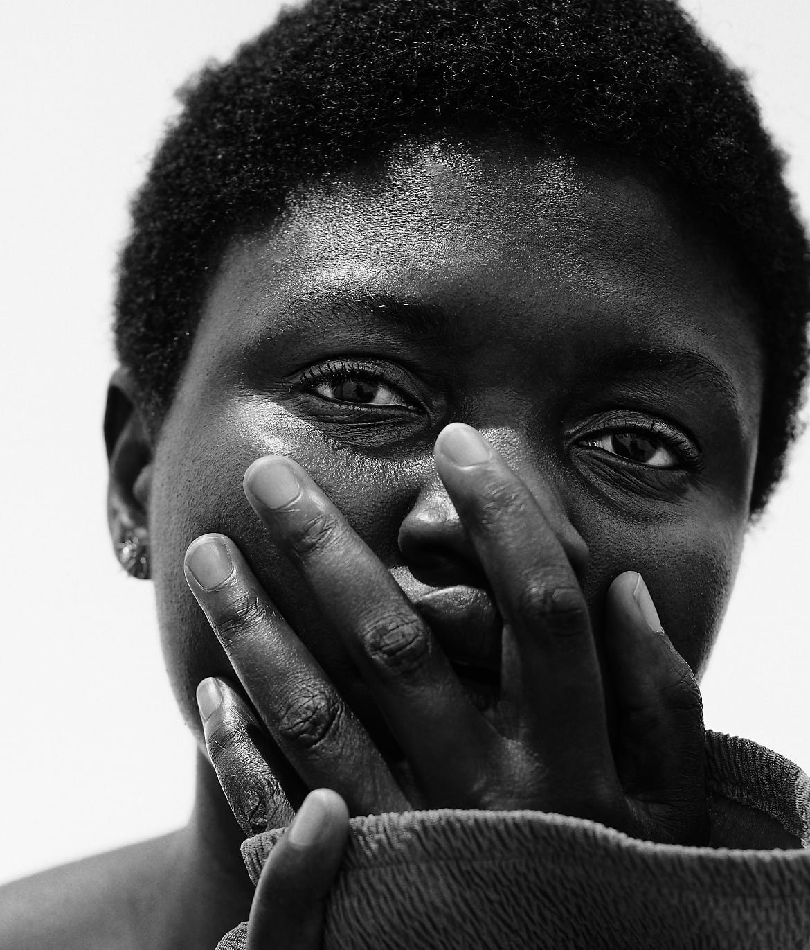 A person with short, natural, tightly coiled hair is shown in a black-and-white portrait. They are wearing a textured garment that covers part of their mouth and hand. The lighting highlights their collarbone and shoulder.