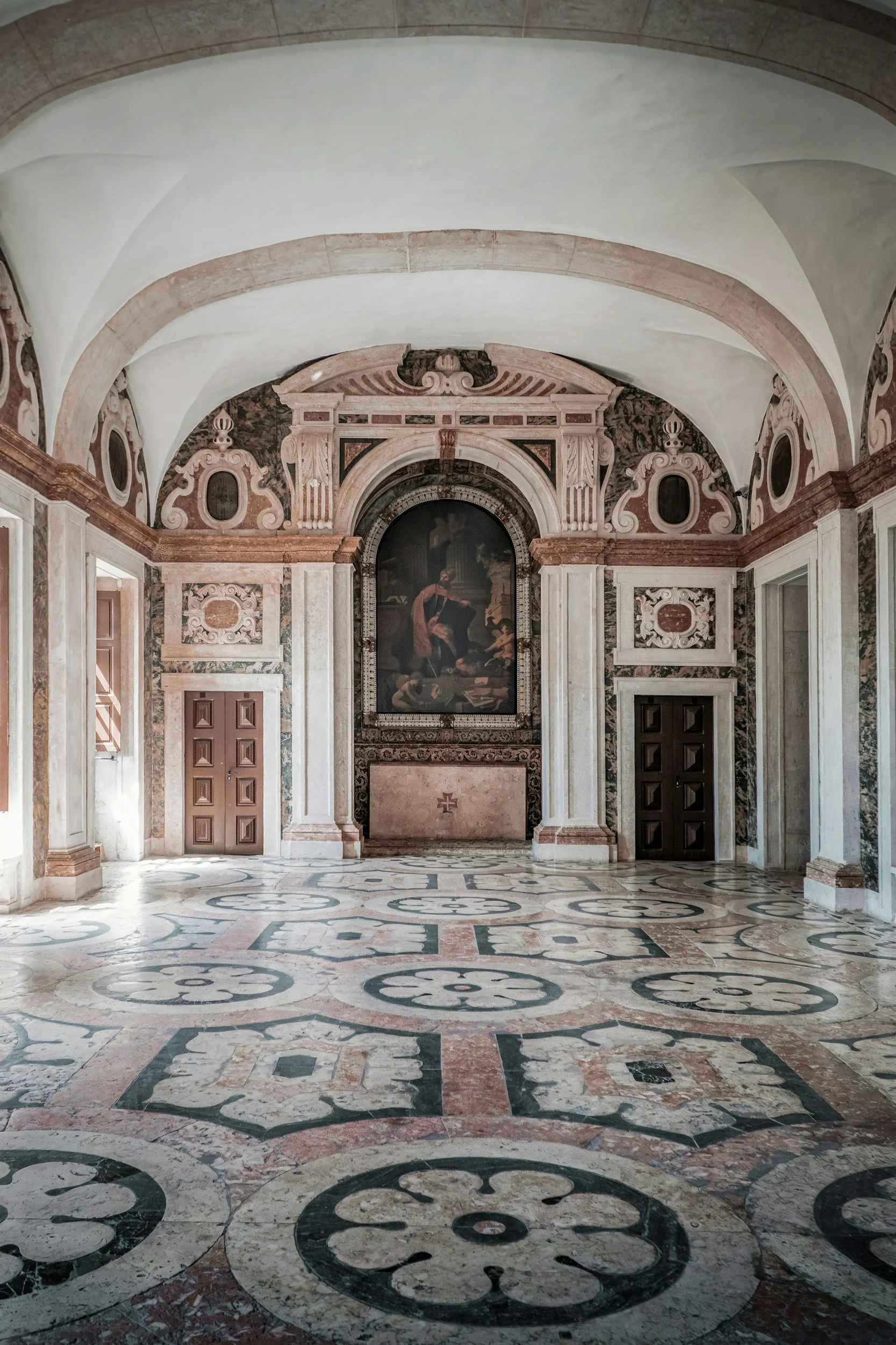 Interior of a historic building with ornate marble walls, decorative moldings, and a large painting of a woman. The floor features intricate marble patterns with circular designs.