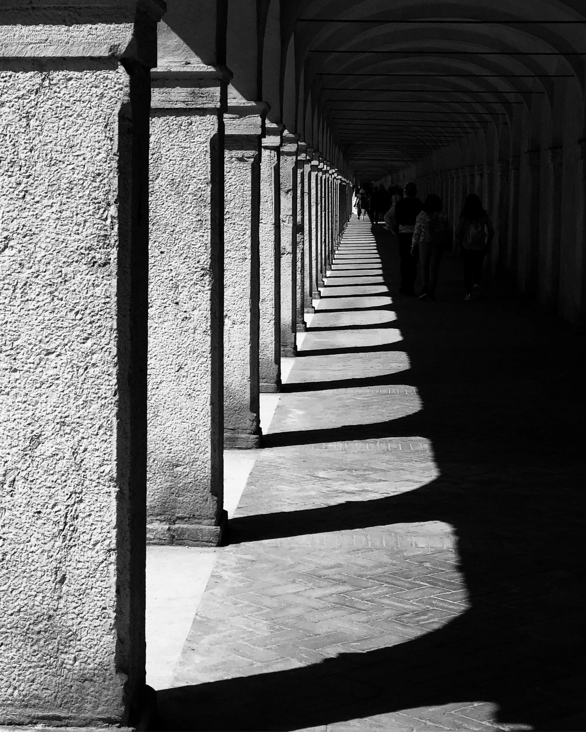Black and white photo of a covered street with stone walls and a row of arches casting shadows on the ground. People are walking in the distance.