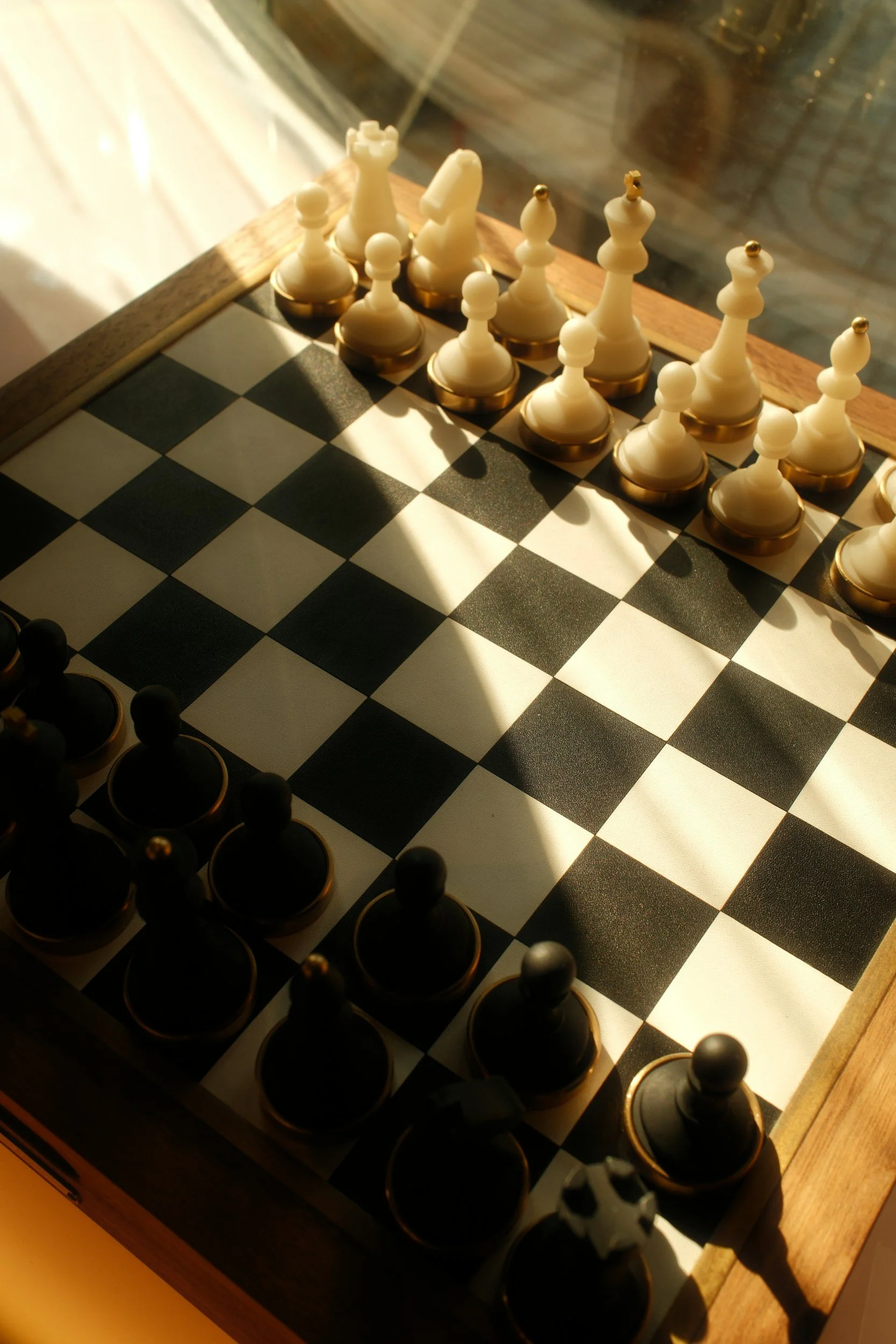 A chessboard with chess pieces set up in starting positions, viewed under warm lighting with a wooden border.
