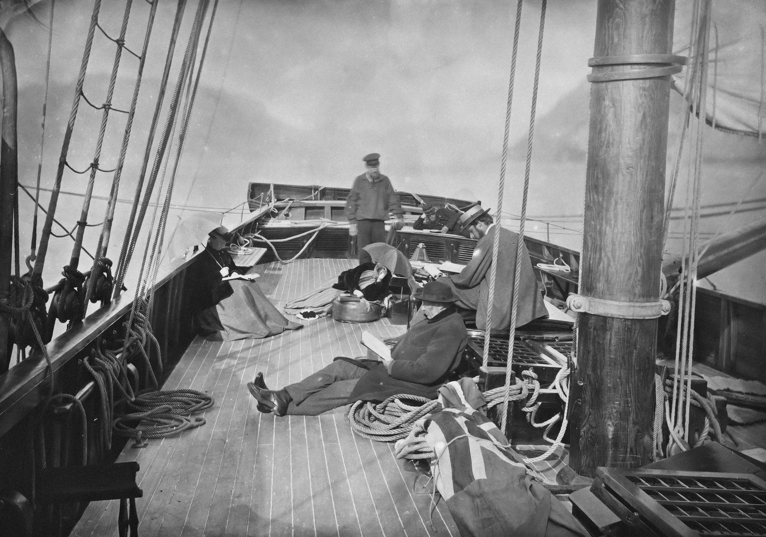 Historical black-and-white photo of the deck of a ship with several men sitting and standing, some reading or writing, with ropes and wooden poles visible. The sky is cloudy.
