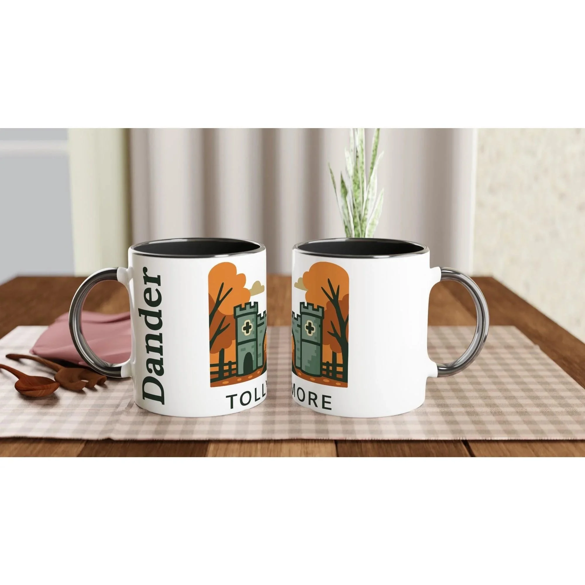 Two white mugs with a castle and trees design, one labeled 'Dander' and the other missing some letters, placed on a table with a checkered placemat and some dried leaves.